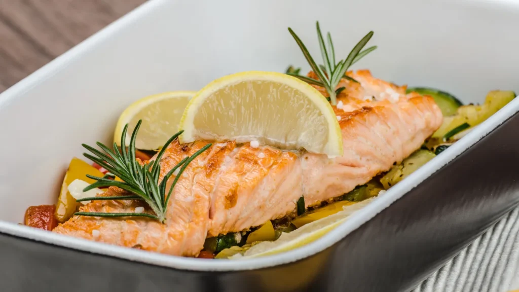 Pavé de saumon cuit au four dans un plat blanc, sur un lit de légumes, avec des rondelles de citron et du romarin